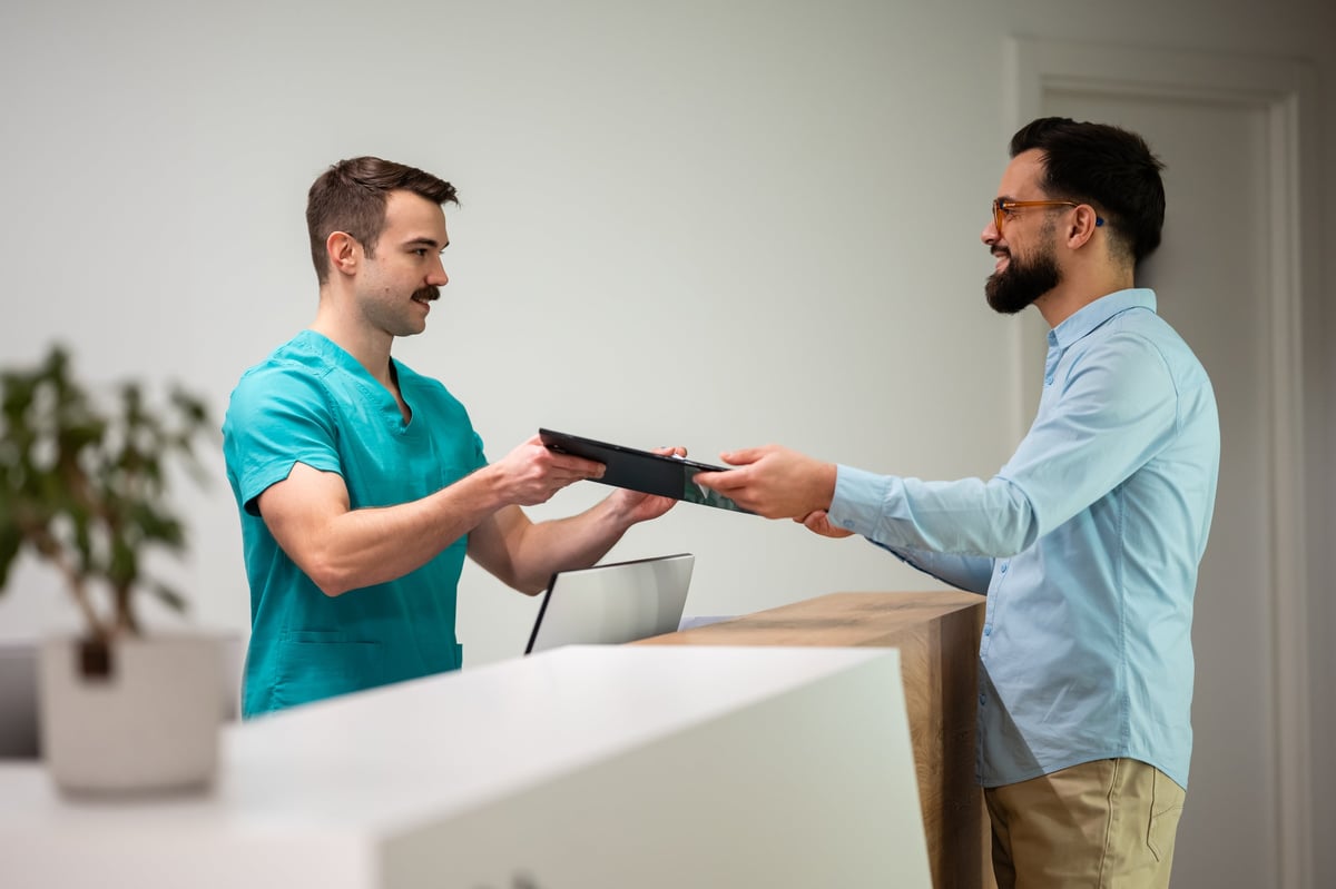 Healthcare worker handing a digital tablet to a patient at a clinic reception desk for registration